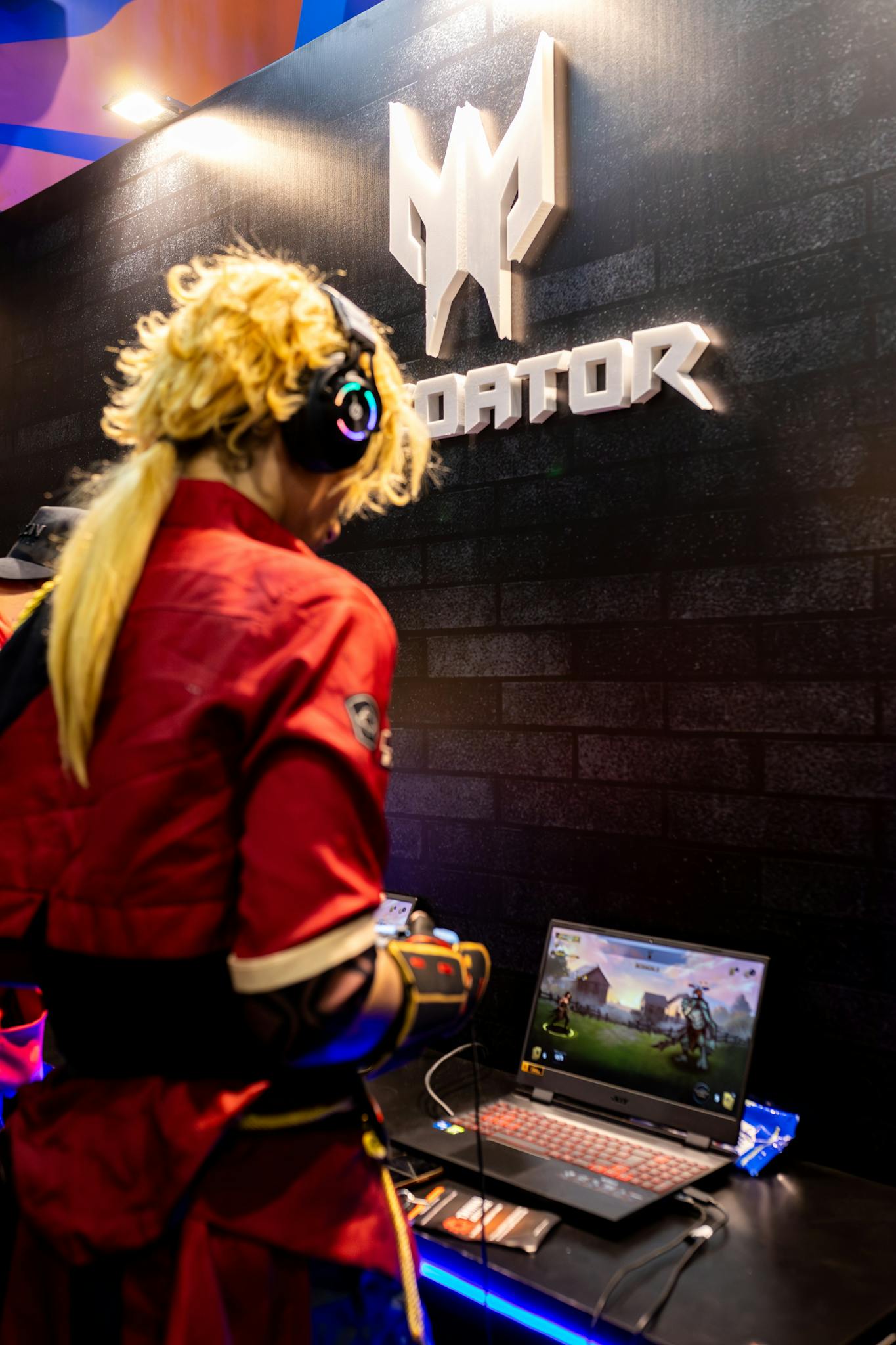 Cosplayer playing a video game at a tech convention booth with gaming laptop.